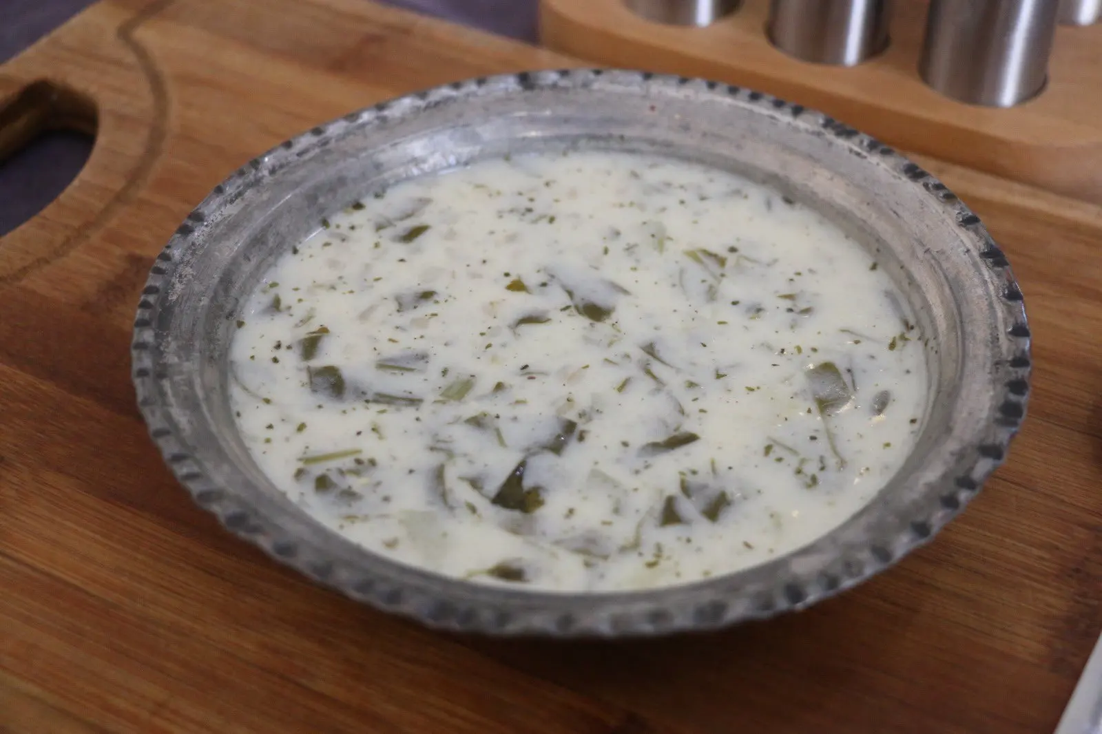 Ahşap bir tepsi üzerindeki metal tabakta, içinde yeşil otlar görünen ayran aşı.