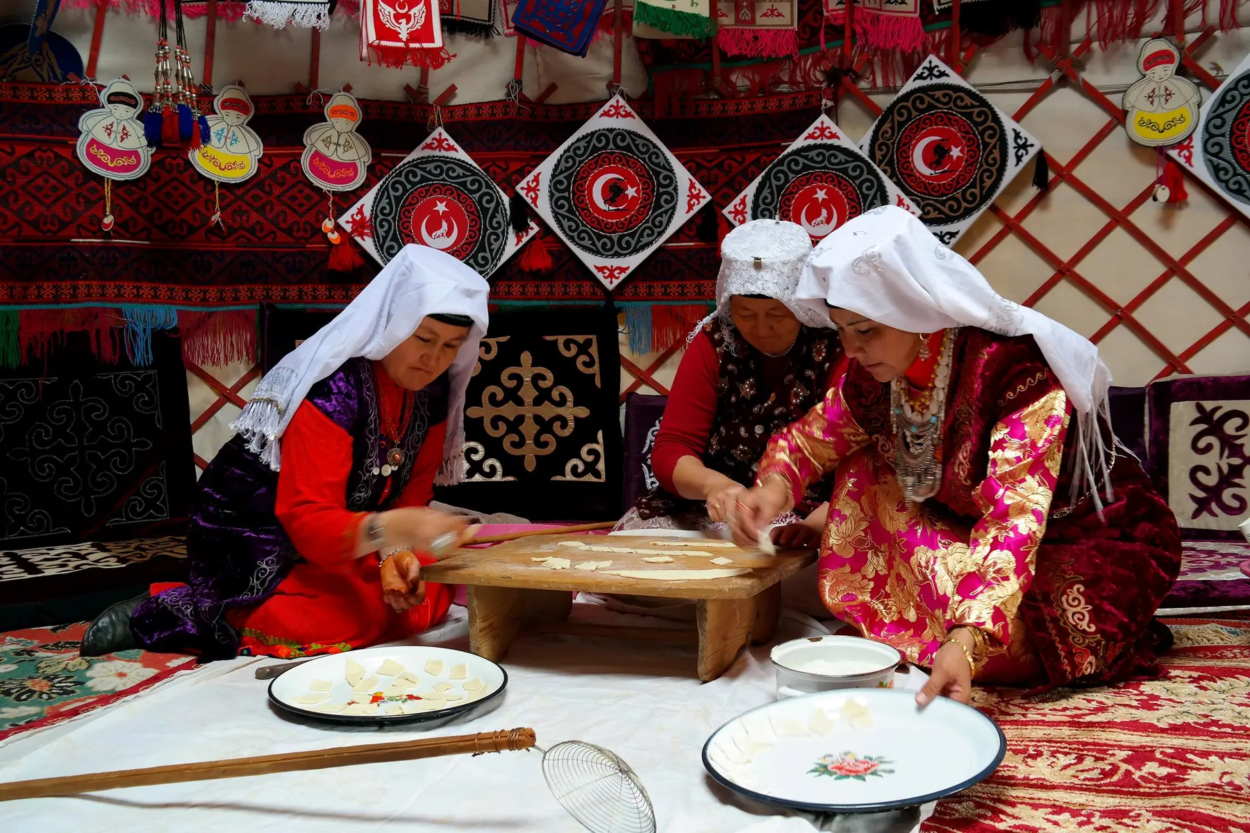 Desenli dokumalarla çevrili bir çadırda, alçak ahşap masa üzerinde hamur hazırlayan üç kadın.